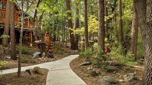 A forested pathway leading to a cabin with a wooden deck and bear sculptures. - Honeycomb Hideaway| Hot Tub & Sauna, Large Yard (Big Bear Lake)