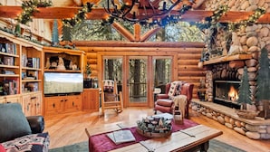 A cozy log cabin interior with a fireplace, bookshelves, a television, and a comfortable seating area. - Honeycomb Hideaway| Hot Tub & Sauna, Large Yard (Big Bear Lake)