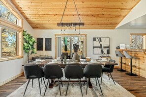A dining area with a wooden ceiling, a large table set for four, and a view through a glass door. - Oak Tree Lodge, w/ Hot Tub, Fire Pit, Game Room (Big Bear)