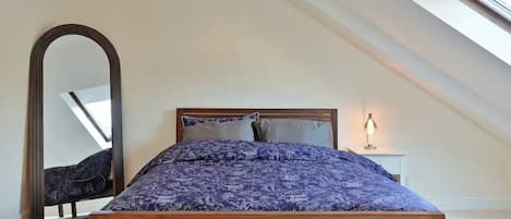 A bedroom with a sloped ceiling, a large bed with a wooden headboard, a bedside table with a lamp, and a full-length mirror.