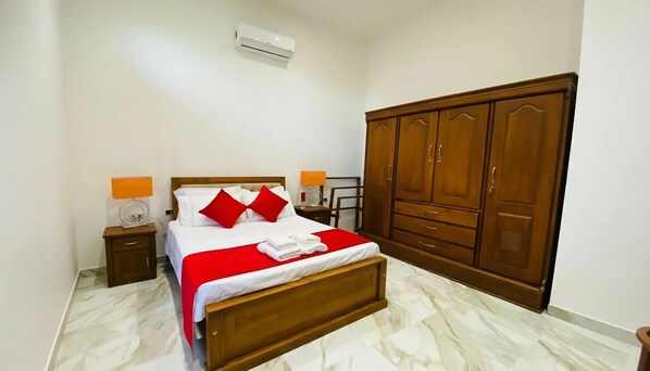 A neatly made bed with red and white bedding, two bedside lamps, a wooden wardrobe, and a marble-patterned floor.