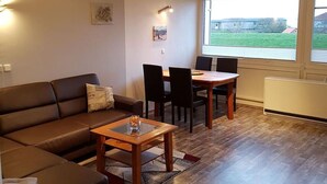 A living room with a sofa, a wooden coffee table, a dining table with chairs, a window with a view of a building, and a radiator. - Apartment 'Meerblick' with Sea View, Balcony and Wi-Fi (Horumersiel-Schillig)