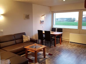 A living room with a sofa, a wooden coffee table, a dining table with chairs, a window with a view of a building, and a radiator. - Apartment 'Meerblick' with Sea View, Balcony and Wi-Fi (Horumersiel-Schillig)