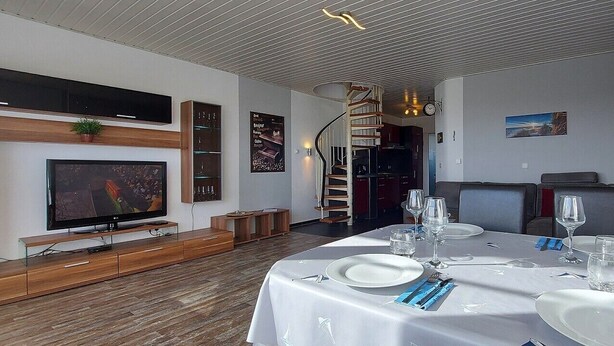 A dining area with a table set for four, a TV on a wooden stand, a staircase, and a wall-mounted shelf with a plant. - Apartment 'Meerblick' with Sea View, Balcony and Wi-Fi (Horumersiel-Schillig)
