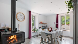A modern kitchen with a built-in wood stove, a dining area with a white table and chairs, and a large window with a view of greenery. - Guest Room “La Drosera - Room No.6” with Shared Terrace, Garden & Wi-Fi (Riols)