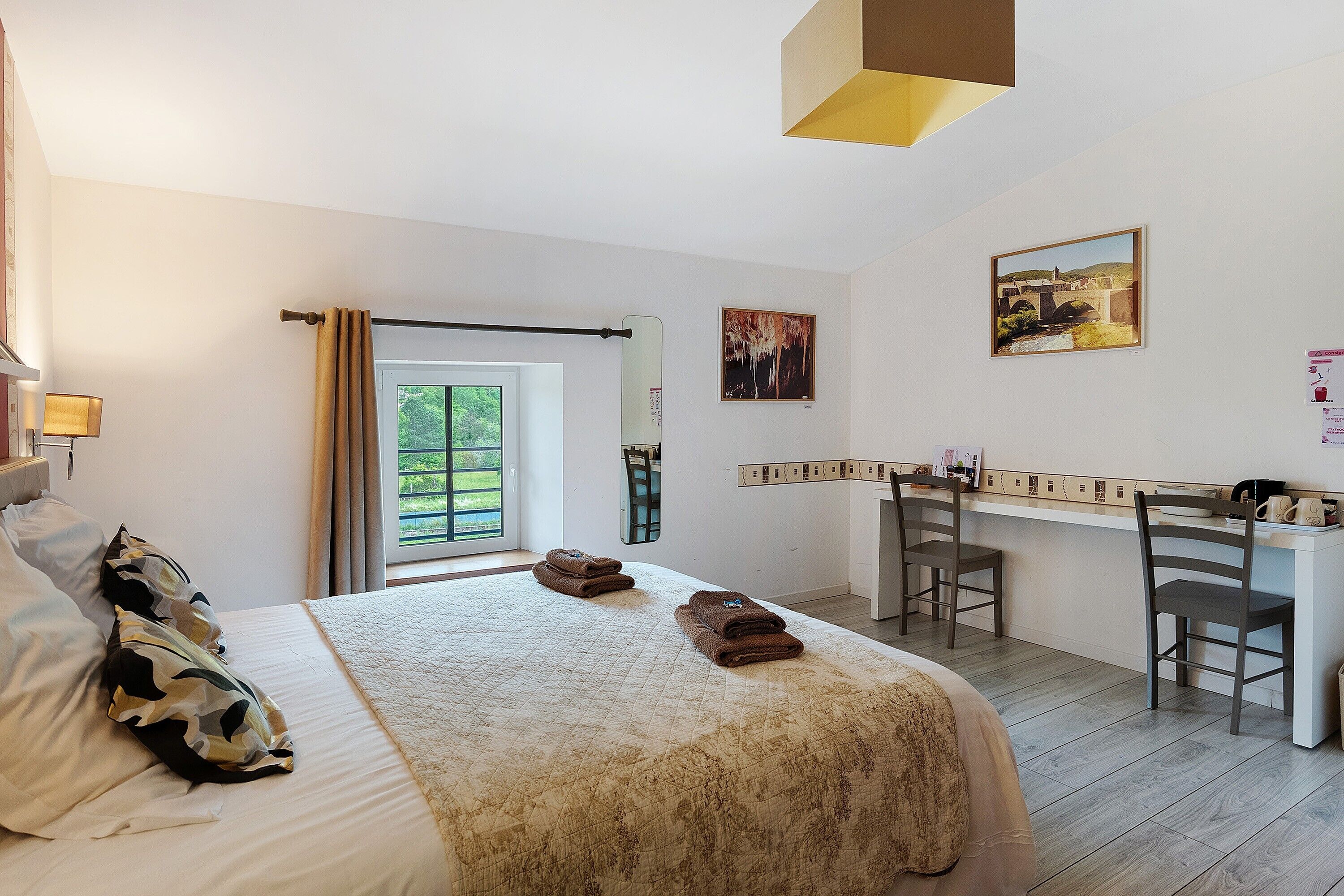 Une chambre d’hôtel comprenant un lit, un bureau, deux chaises, une fenêtre avec des rideaux et des œuvres d’art accrochées aux murs.