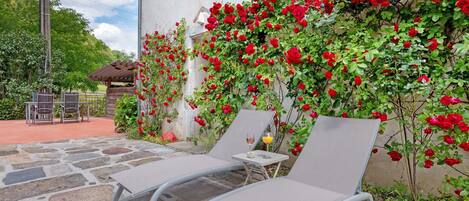 Un patio de piedra con dos sillones, una mesita con un vaso de zumo de naranja y una pared cubierta de rosas rojas.