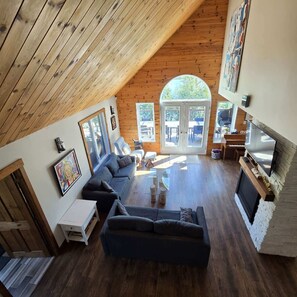 A cozy living room with a sloped ceiling, a fireplace, and a large window overlooking a deck.