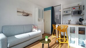 Una sala de estar moderna con un sofá gris, una mesita redonda con una planta, y una cocina con microondas, armarios y cafetera.