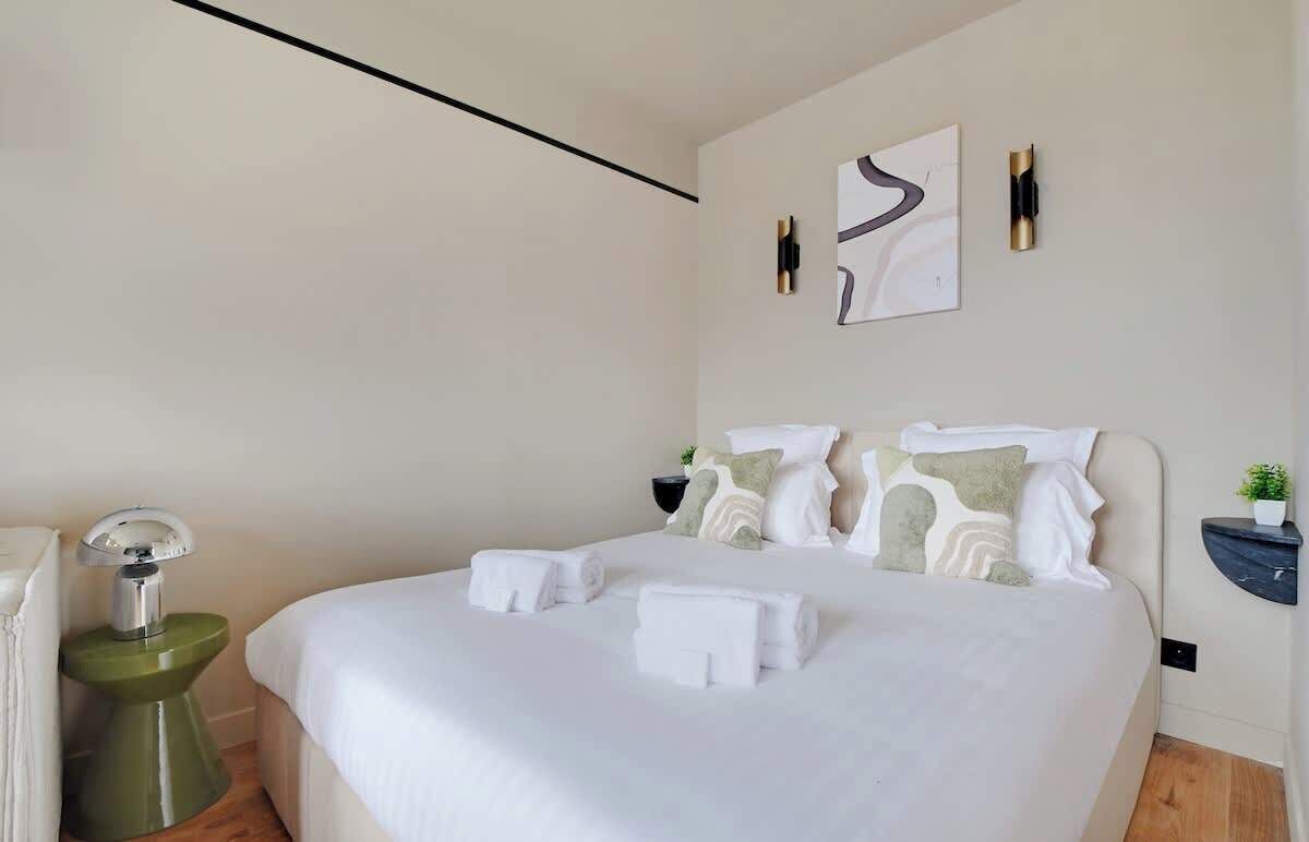A neatly made bed with white linens and towels, a bedside table with a lamp, and a wall-mounted artwork.