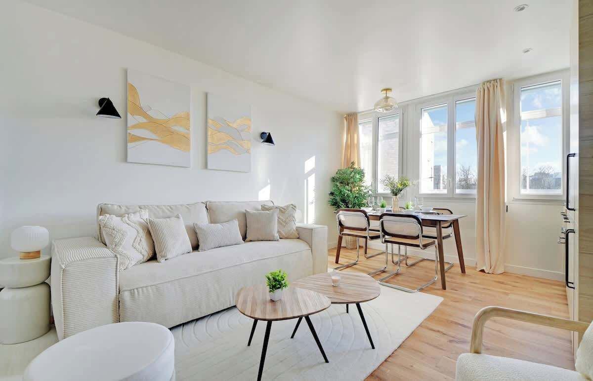 A modern living room with a sofa, coffee table, and dining area. There are two paintings on the wall, a plant, and a window with curtains.