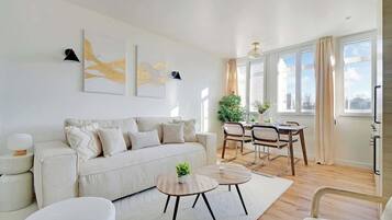 A modern living room with a sofa, coffee table, and dining area. There are two paintings on the wall, a plant, and a window with curtains.
