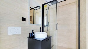 Un baño moderno con ducha de vidrio, tocador negro y lavamanos blanco, y un espejo sobre el lavamanos.