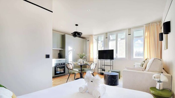Habitación de hotel moderna con cama, zona de comedor, televisor y cocina al fondo.