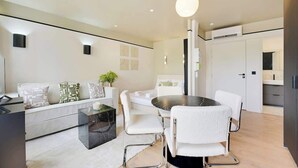 Ein modernes Wohnzimmer mit einer Couch, einem Couchtisch und einem Esstisch mit Stühlen. Auf der Couch steht eine Pflanze und über dem Esstisch hängt eine Pendelleuchte.