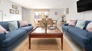 Ein Wohnzimmer mit zwei blauen Sofas, einem Holztisch, einem Esstisch mit Stühlen und einem an der Wand montierten Fernseher.