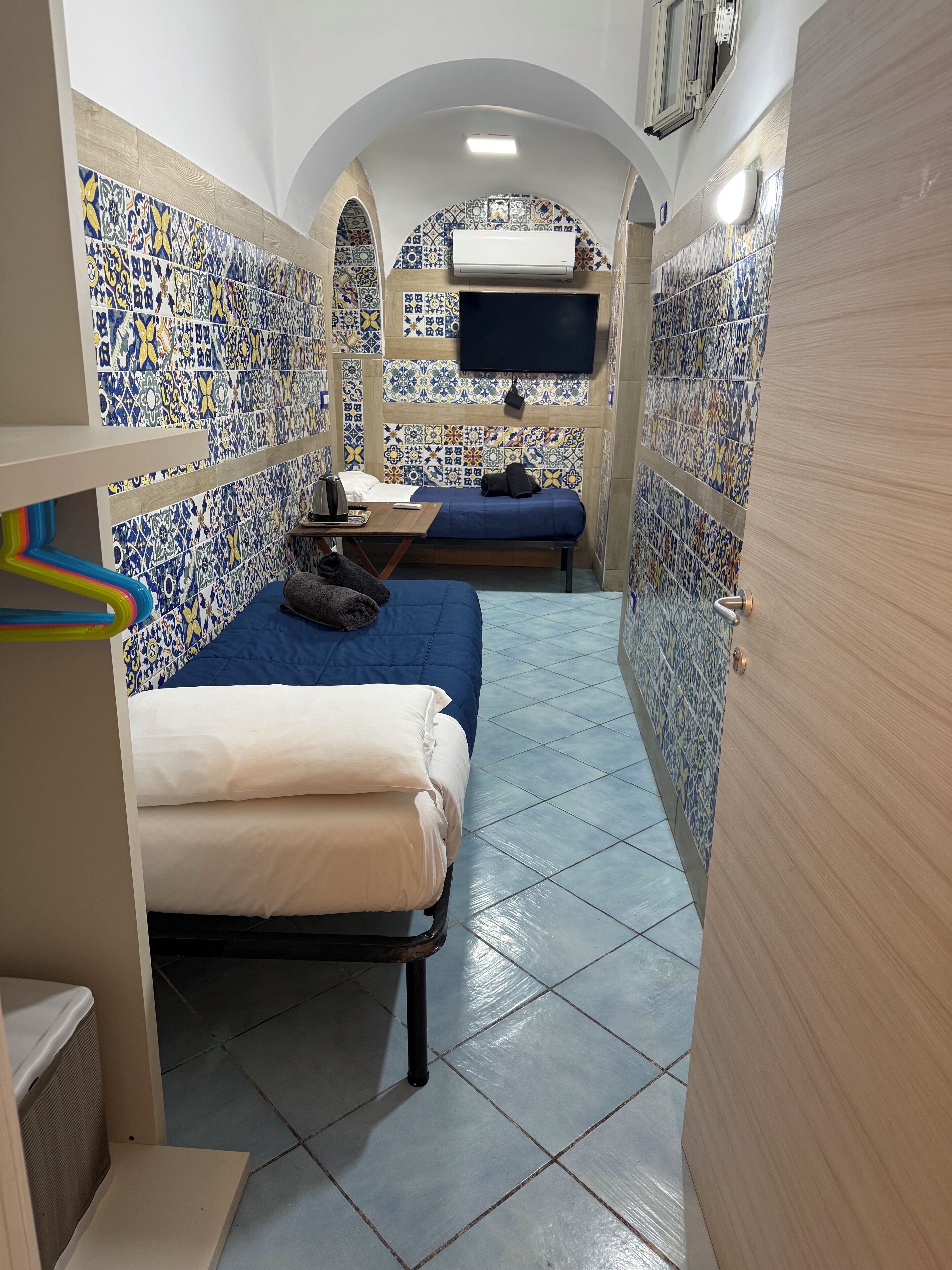 A narrow room with two beds, a TV mounted on the wall, and tiled walls with a blue and white pattern.