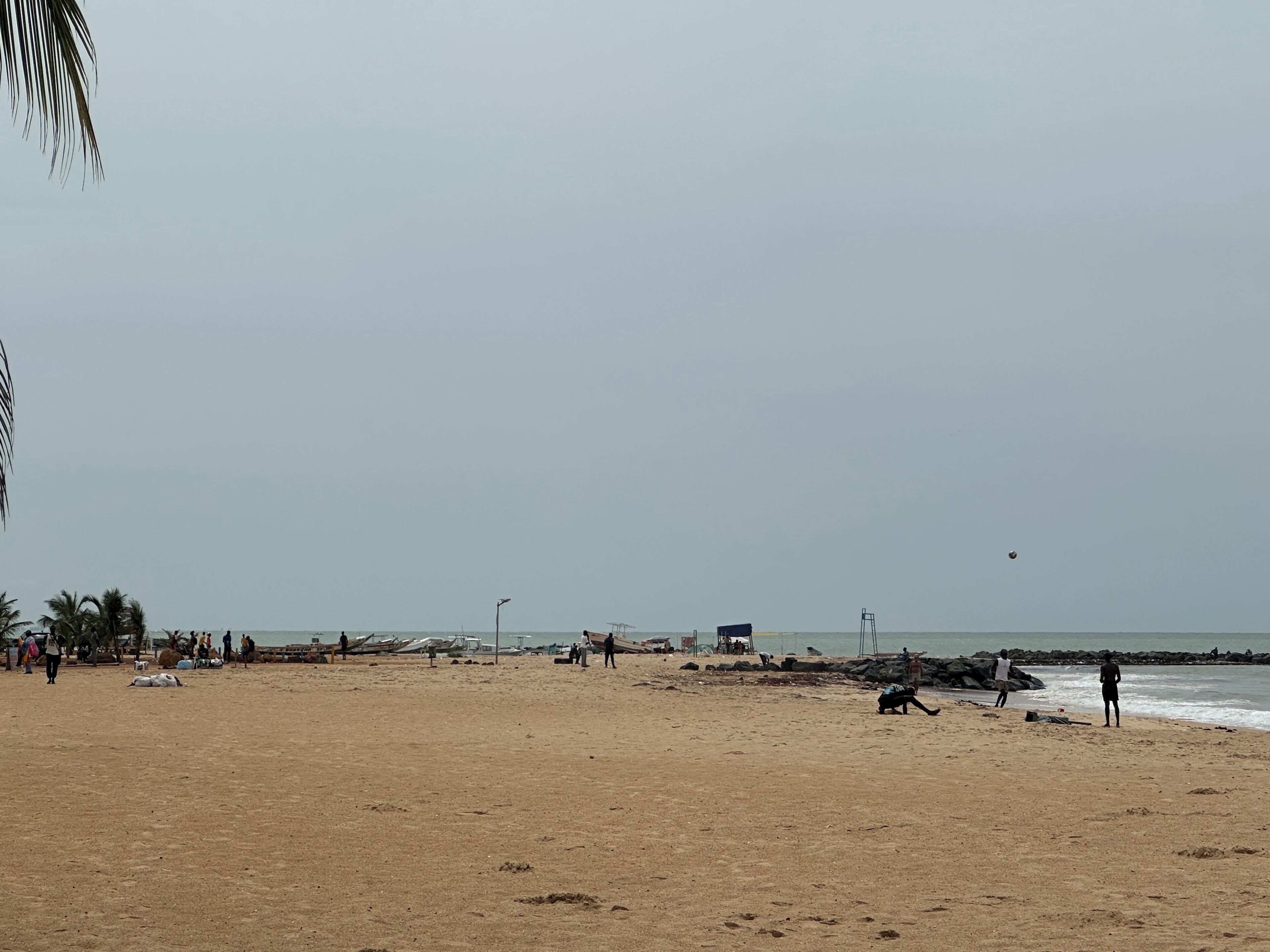 Een breed zandstrand met enkele mensen verspreid over het strand, sommigen dichtbij het water en anderen op het zand. Er staan een paar gebouwen en een lantaarnpaal in de verte.