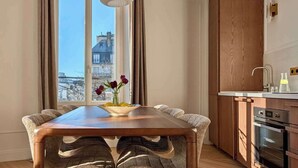 A dining area with a wooden table and chairs, a vase with flowers, and a view of buildings through large windows. - A Charming 2-BR/2BA - Montorgueil (Paris)