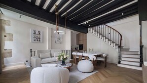 A modern living room with a sofa, ottoman, and dining table. A staircase with a railing is visible in the background. - An Exceptional 3-BR/3Ba with Services - Le Marais (Paris)