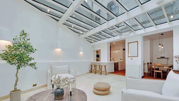 A spacious living area with a high ceiling and skylight, a round wooden coffee table, white sofas, and a dining area in the background. - A Breathtaking 2-BR/1Ba - Gobelins (Paris)