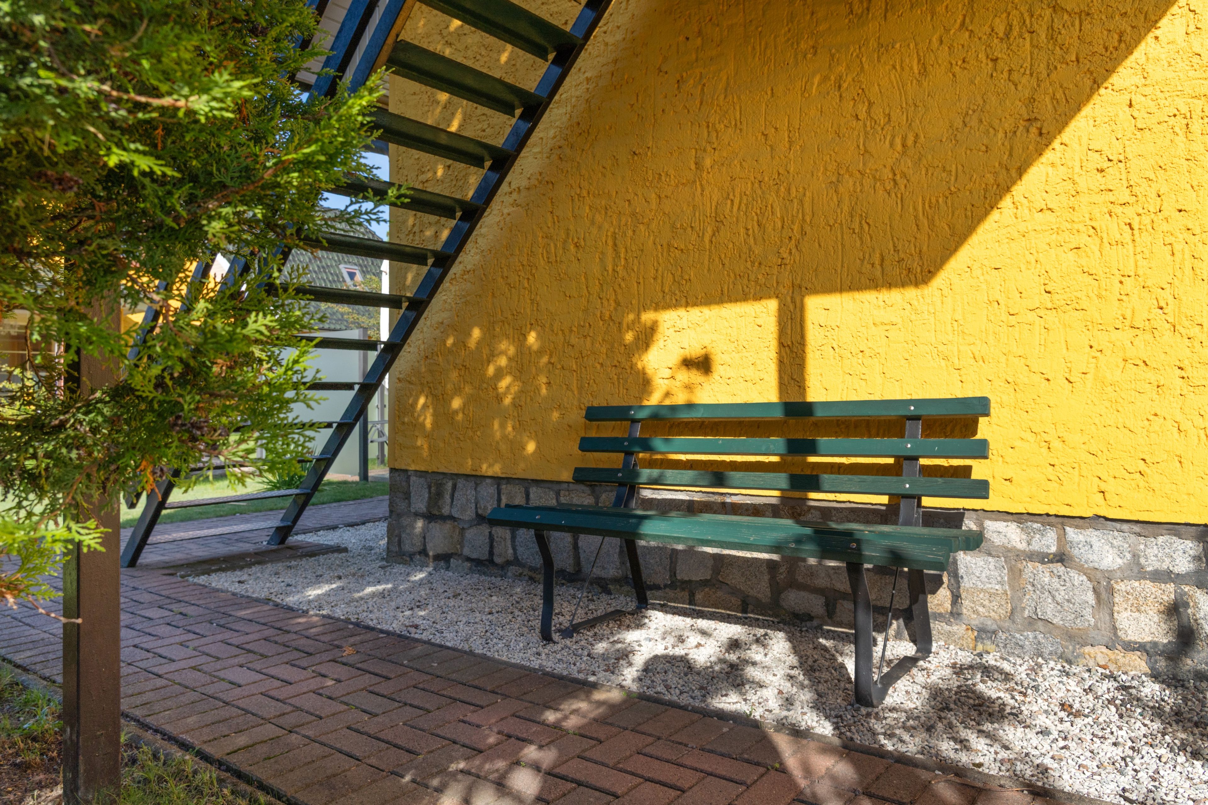 A bench against a yellow wall with a stone base, a metal ladder, and a tree in the foreground.