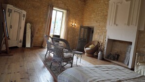 A rustic room with a wooden floor, a bed with a quilt, wicker chairs, a fireplace, and a stone wall. - Château de Saint Chamarand (Saint-Chamarand)