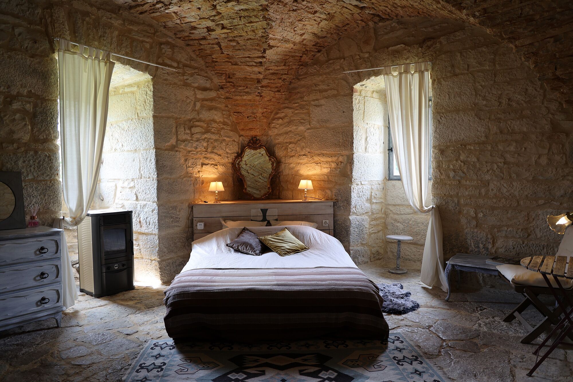 A stone-walled bedroom with a bed, two bedside lamps, a dresser, and a mirror on the wall.