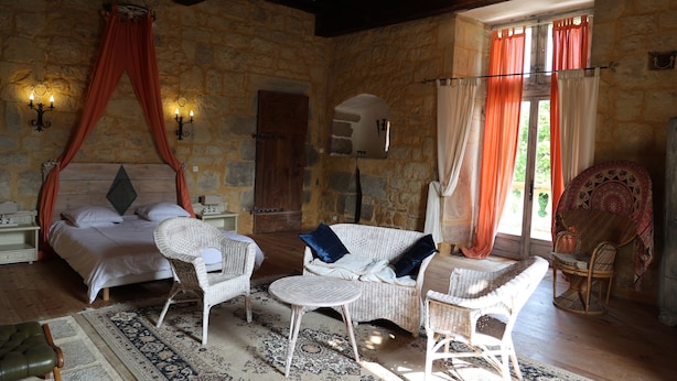 A bedroom with a stone wall, a canopy bed, wicker furniture, and a large window with curtains. - Château de Saint Chamarand (Saint-Chamarand)