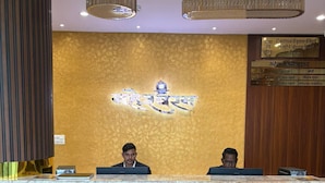 A reception desk with two employees, a decorative wall with a logo, and a plaque with text. - Hotel Maheshwaram Vijay Nagar Indore (Indore)