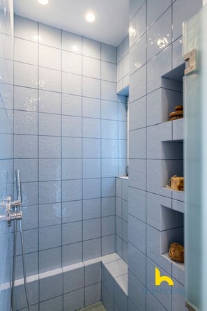 A bathroom with blue tiles, a shower area, and built-in shelves with toiletries. - Stylish Love Nest W/ Balcony in Heart of Koukaki! (Athina)