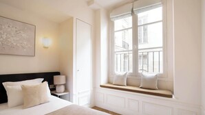Een hotelkamer met een groot bed, een nachtkastje met een lamp, een raam met uitzicht op gebouwen en een schilderij aan de muur.