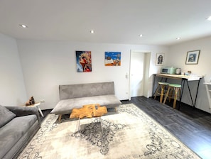 A modern living room with a grey sofa, a coffee table, and a patterned rug. There are two wall art pieces, a door, and a small kitchen area with a microwave. - The High Gallery: An exclusive 230 m² apartment designed by an artist in the heart of Hirschaid (Hirschaid)