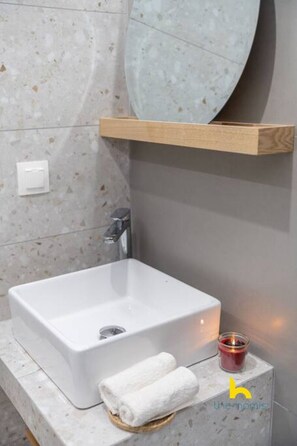 A bathroom with a white sink, a wooden shelf above it, and a small candle on the counter. - Kypseli's Dreamy Studio Your Cosy Greek Oasis! (Athina)