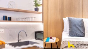 A modern kitchen with a sink, stove, and microwave. There is a small table with a book and a candle. The room has wooden paneling and a bed with pillows. - Kypseli's Dreamy Studio Your Cosy Greek Oasis! (Athina)