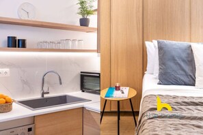 A modern kitchen with a sink, stove, and microwave. There is a small table with a book and a candle. The room has wooden paneling and a bed with pillows. - Kypseli's Dreamy Studio Your Cosy Greek Oasis! (Athina)
