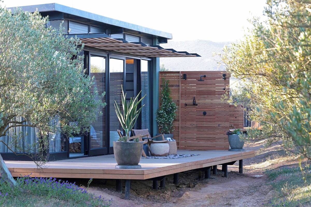 Une maison moderne dotée d’une terrasse en bois, de grands pots de plantes et d’un portail en métal.