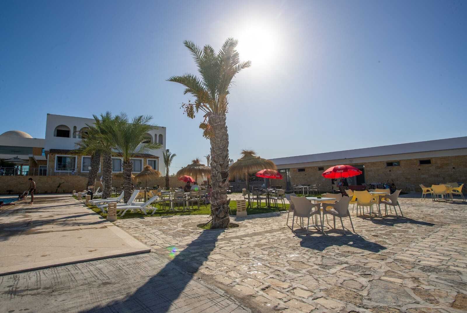 A sunny outdoor area with palm trees, outdoor seating, and a building with a dome and arched windows.