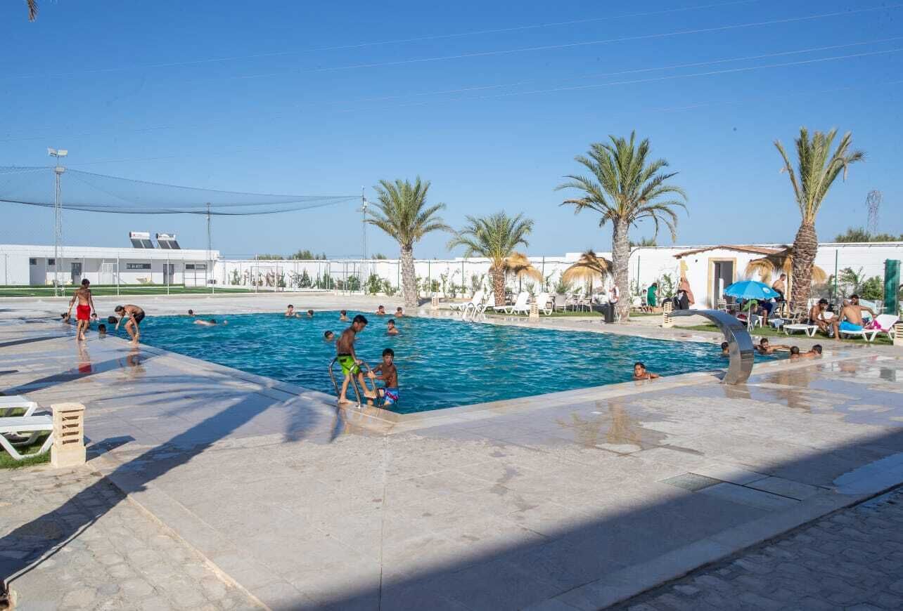 Una piscina con gente alrededor, palmeras y un edificio al fondo.