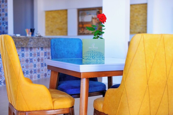 A dining table with a vase of a red rose, two blue chairs, and a yellow chair. - DAR AMMAR (El Djem)
