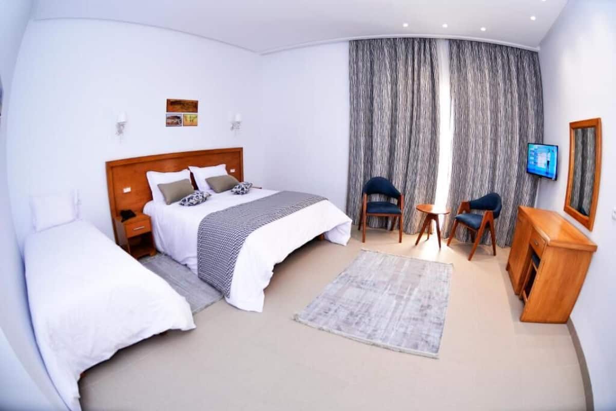 A modern hotel room with a large bed, two bedside tables, a small desk with chairs, a TV, and a large window with curtains.