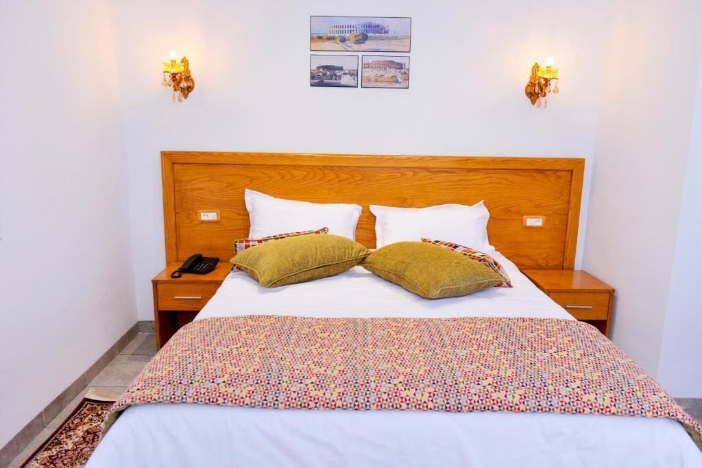 Una cama bien tendida con una colcha estampada, dos mesitas de noche con lámparas y un cuadro colgado en la pared.