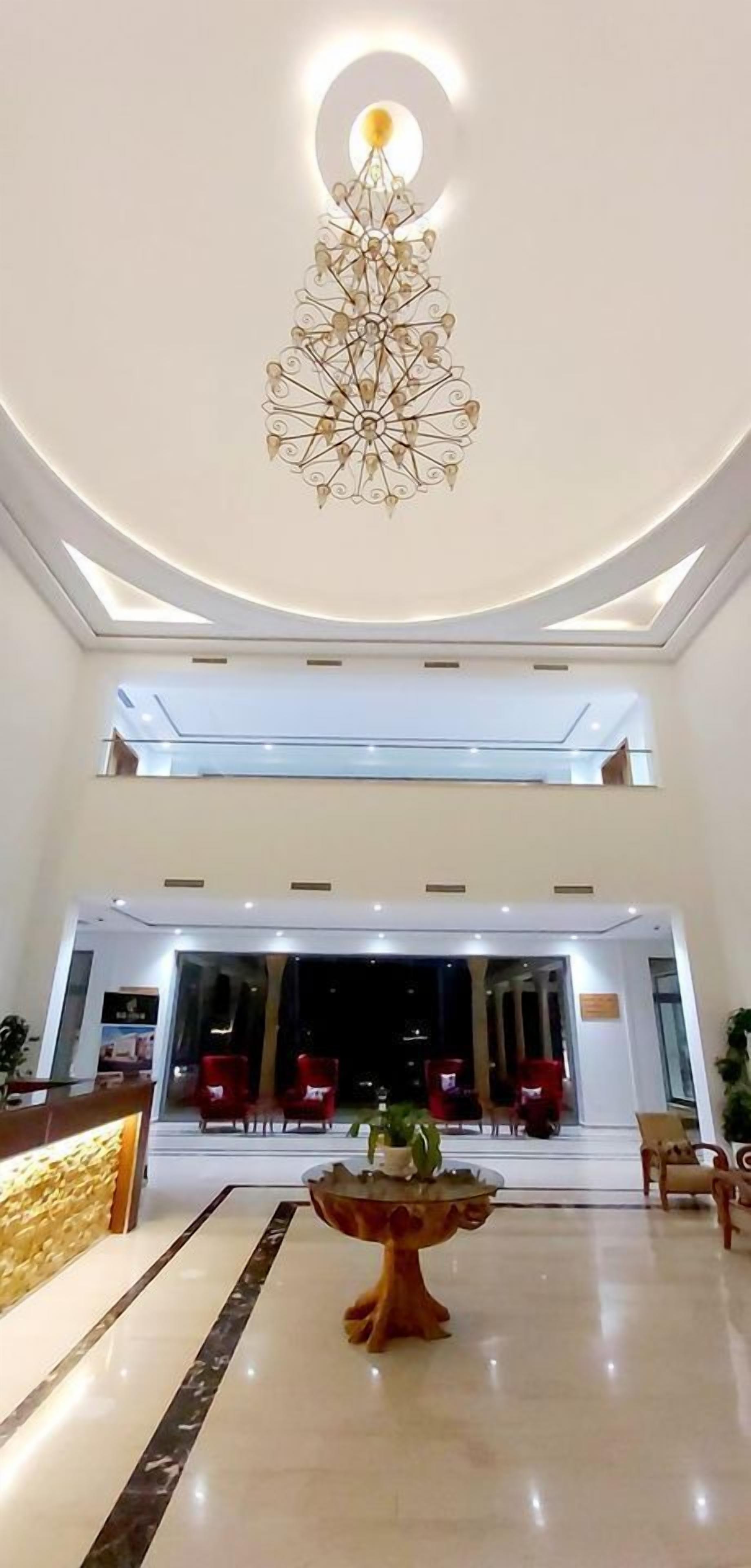 A hotel lobby with a modern chandelier, a reception desk, and red chairs.