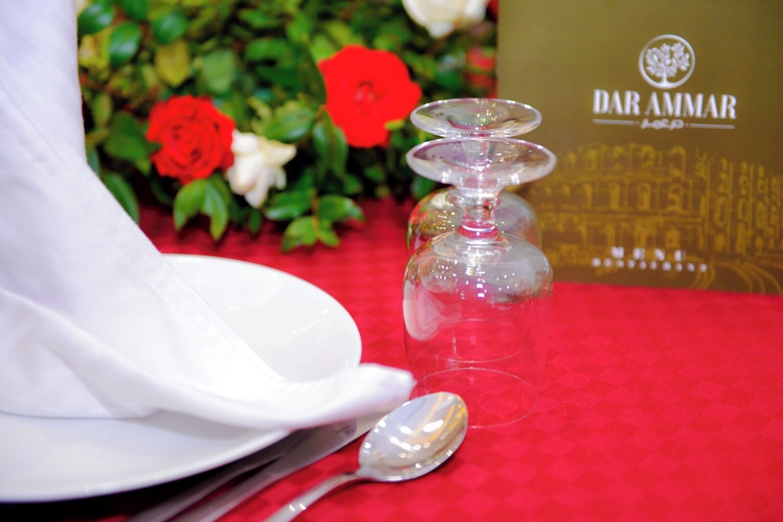 Une table dressée avec une serviette blanche, un verre à vin transparent et une cuillère posés sur une nappe rouge.