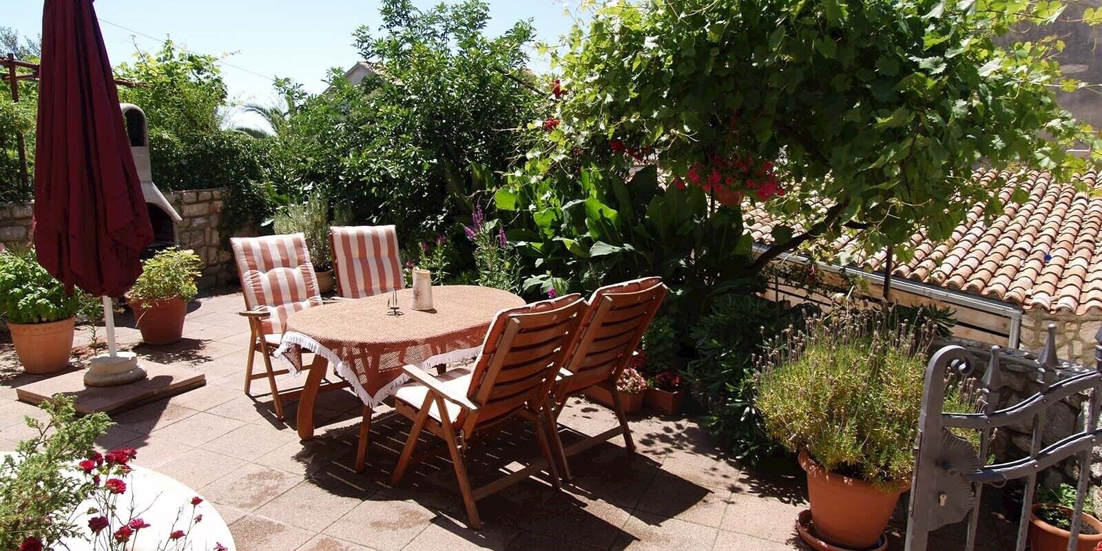 Une terrasse extérieure avec une table ronde et des chaises, entourée de plantes en pot et d’un parasol rouge.