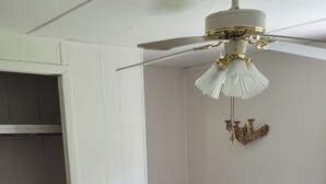Ein Zimmer mit Deckenventilator, ein Holzstuhl mit Kissen, einem Bett mit Steppdecke und einem Kleiderschrank.
