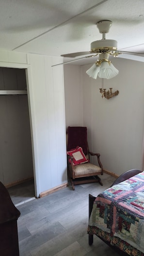 Ein Zimmer mit Deckenventilator, ein Holzstuhl mit Kissen, einem Bett mit Steppdecke und einem Kleiderschrank.