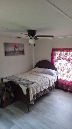 A single bed with a headboard, a ceiling fan, a painting of the Eiffel Tower, and a window with floral curtains.