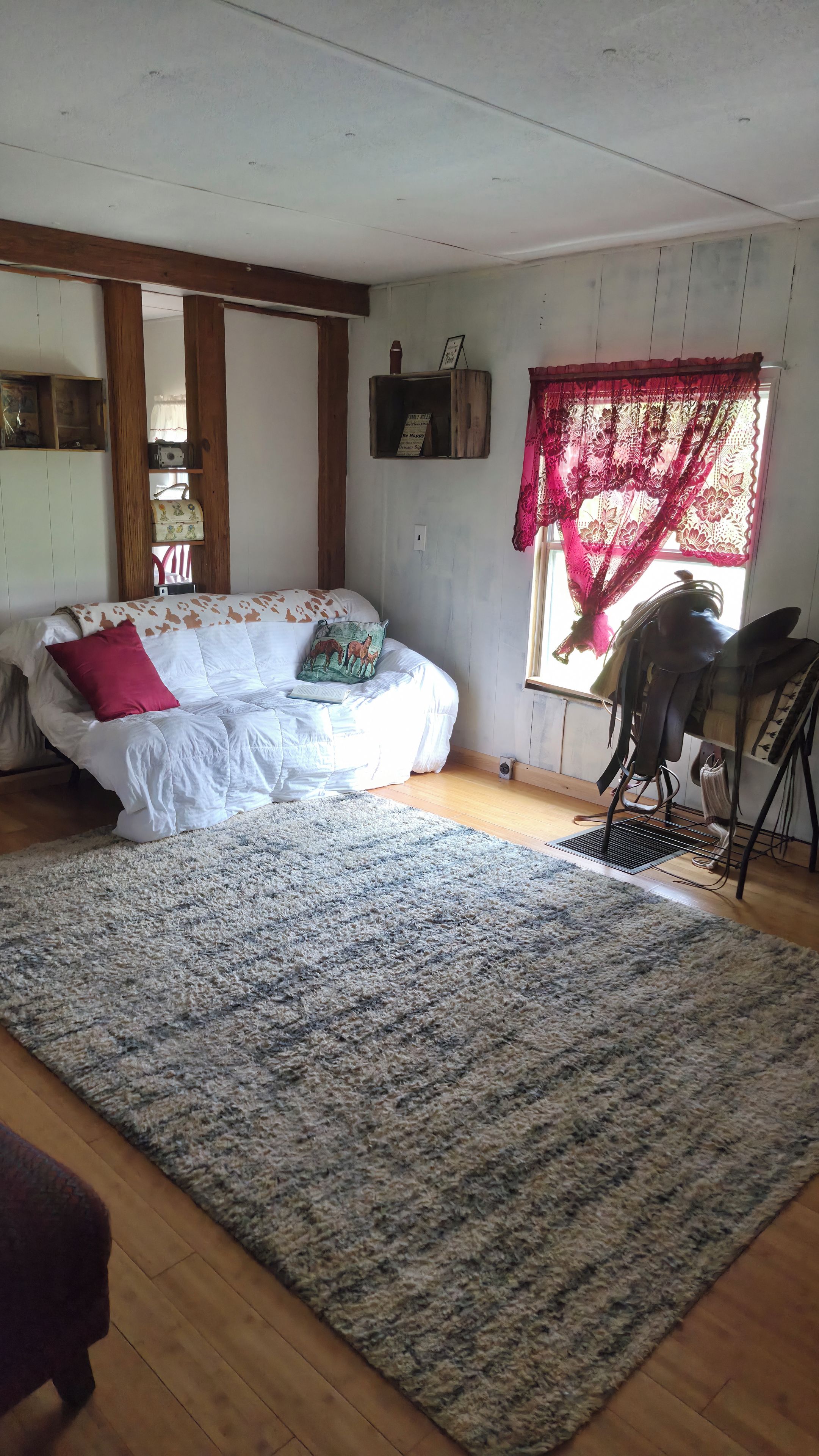 A room with a white sofa, a patterned rug, wooden flooring, a window with lace curtains, and a wall-mounted shelf with a framed picture.