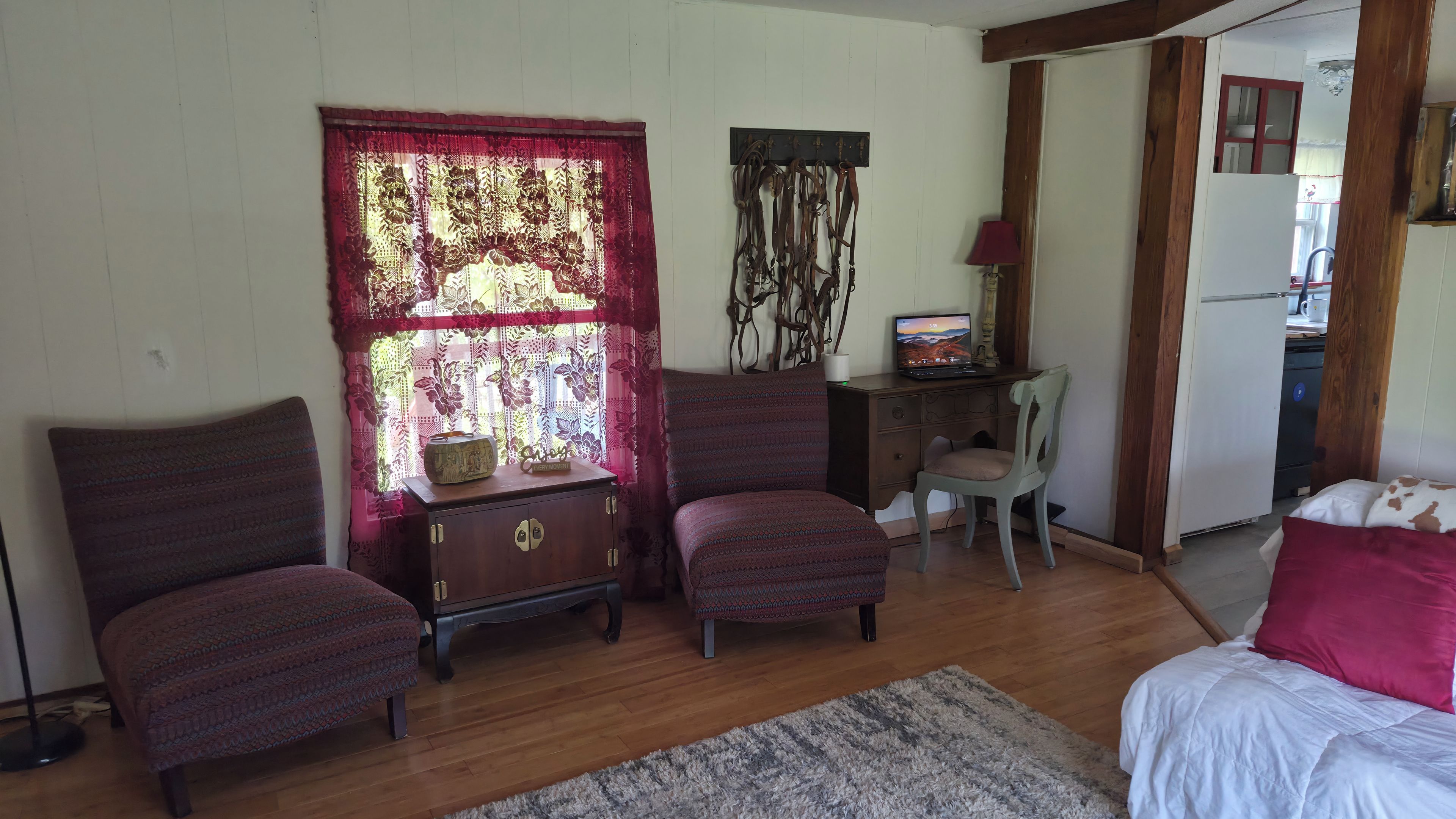 A room with two armchairs, a small wooden cabinet, a desk with a computer, a chair, a bed with a white and brown blanket, and a window with a red curtain.